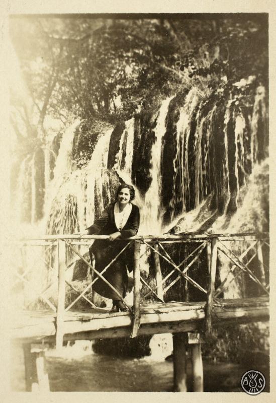 Retrat de dona sobre un pont de fusta amb un salt d'aigua al fons. S.l.