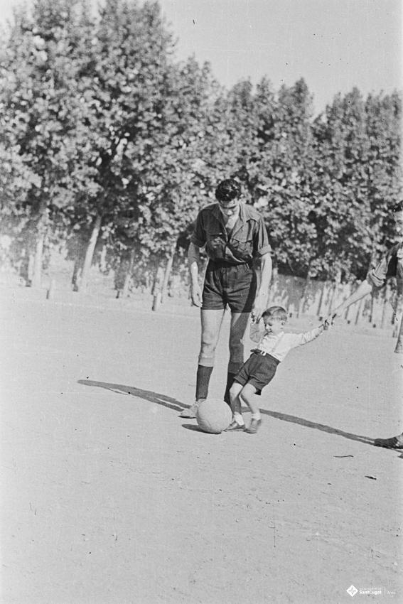 Un infant xuta una pilota de futbol de la mà de dos futbolistes, a l'antic camp de futbol situat al recinte del Monestir.