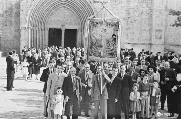 Actes de la festa inaugural de la restaurada Societat Coral La Unió Santcugatenca. Pere Rifé portant l'estendard de la Unió encapçala un grup de gent a la sortida de missa, a la llotgeta del Monestir.