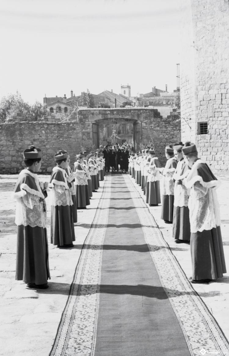 Dues fileres d'escolans esperen l'entrada a la llotgeta d'un grup de persones que just travessen la porta del recinte de l'església de Sant Pere d'Octavià.