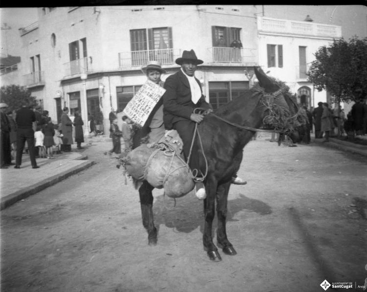 Festa de Sant Antoni Abat. Retrat d'un home i un infant a lloms d'un ruc a la plaça de Lluís Millet. L'infant du un cartell a les mans.