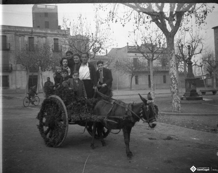 Festa de Sant Antoni Abat. Retrat d'un grup de persones en un carro guarnit,  just davant del brollador de benzina de la plaça d'Octavià. Al fons de la imatge és veu l'edifici de Cal Boter Negre.