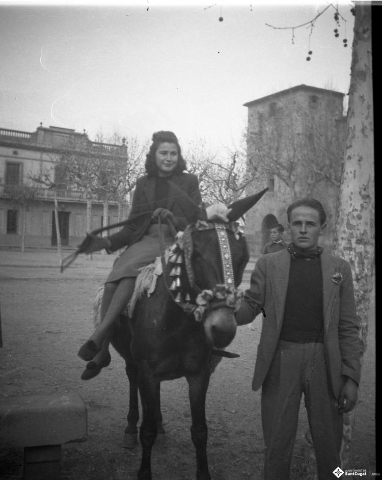 Festa de Sant Antoni Abat. Retrat d'una noia jove muntant un cavall portat per un home, al fons Cal Boter Negre, a la plaça d'Octavià.