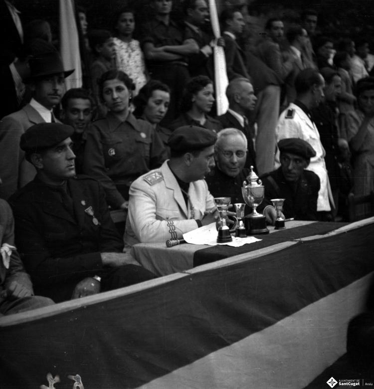 Festa Major. Autoritats assegudes darrere una taula amb copes i trofeus, presideixen l'acte, el governador civil i Antonio Federico Correa Véglison, mossèn Griera,  i Nicolau Villar Recio.