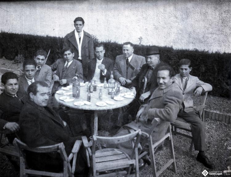 Celebració davant de la seu de la Falange a Cal Barbany. Retrat d'un grup d'homes en una de les taules del quiosc de begudes de la plaça del Quatre Cantons.