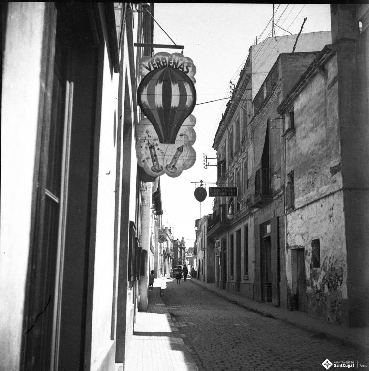 Carrer de Santa Maria, en primer terme, un rètol de l'establiement  dels Cabanas on es pot llegir "Verbenas".
