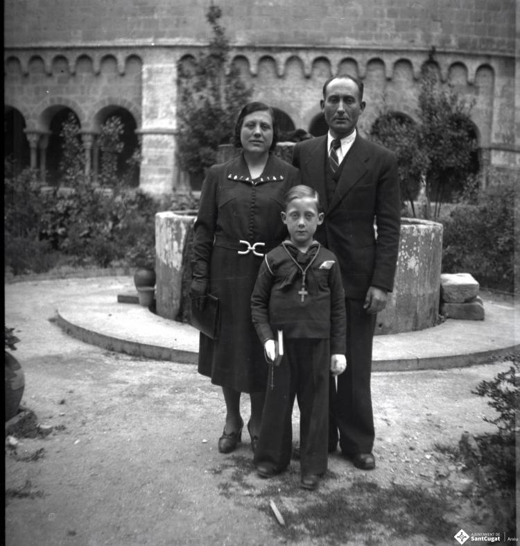 Retrats d'un infant, vestit per a la primera comunció, amb els seus pares davant de la font del pati del claustre del Monestir de Sant Cugat del Vallès.