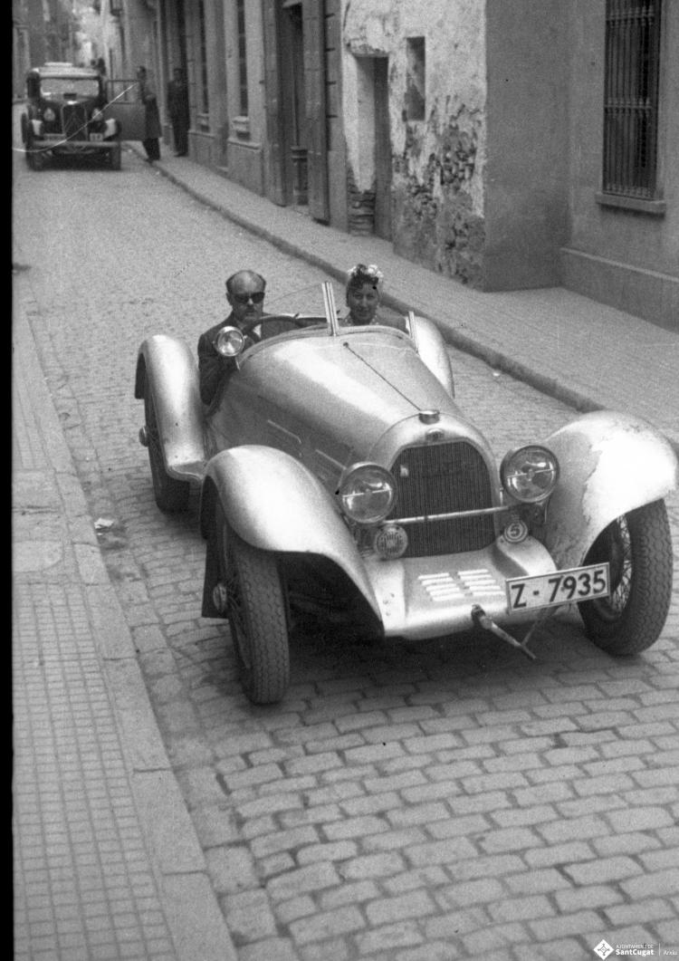 Vehicles circulant pel carrer de Santa Maria, en primer terme una parella en un automòbil esportiu descapotable.