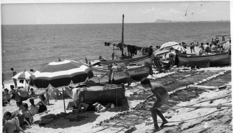 Banyistes prenent el sol entre les barques i les xarxes de pesca