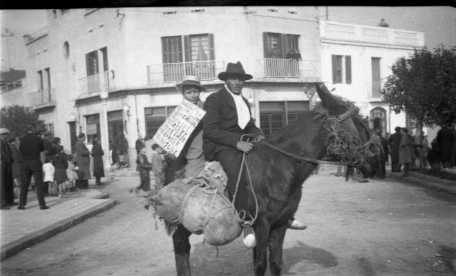 Festa de Sant Antoni Abat. Retrat d'un home i un infant a lloms d'un ruc a la plaça de Lluís Millet. L'infant du un cartell a les mans.