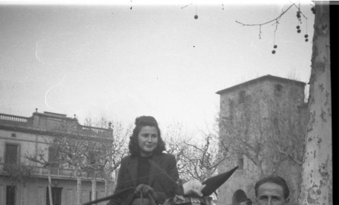 Festa de Sant Antoni Abat. Retrat d'una noia jove muntant un cavall portat per un home, al fons Cal Boter Negre, a la plaça d'Octavià.