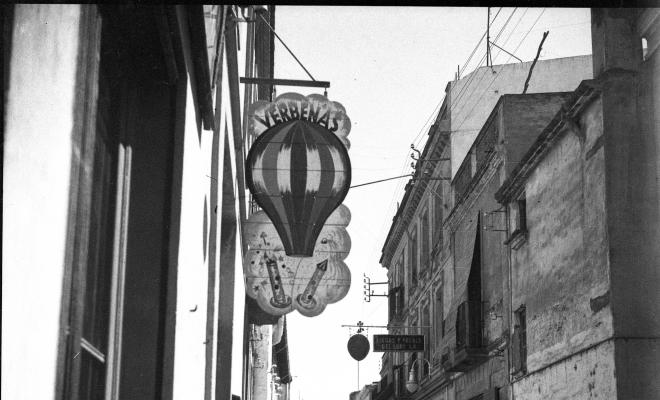 Carrer de Santa Maria, en primer terme, un rètol de l'establiement  dels Cabanas on es pot llegir "Verbenas".