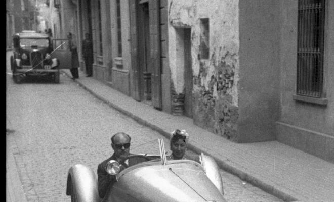 Vehicles circulant pel carrer de Santa Maria, en primer terme una parella en un automòbil esportiu descapotable.